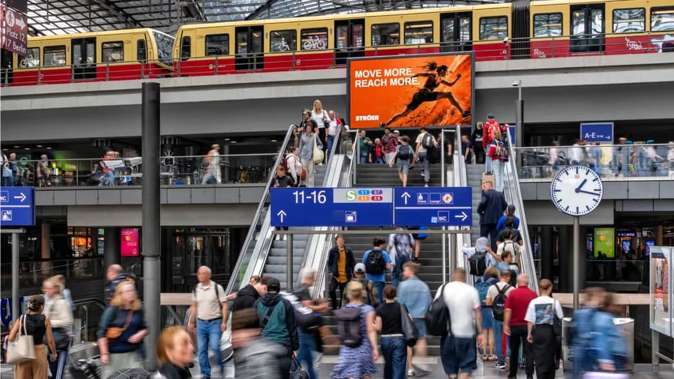 Menschen bewegen sich durch einen Bahnhof, im Hintergrund ist ein großer digitaler Werbescreen zu sehen.