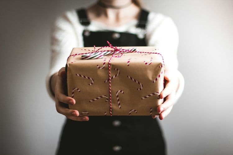 Person hält ein in braunes Papier verpacktes Paket mit rot-weißer Schnur nach vorn; Hintergrund unscharf.