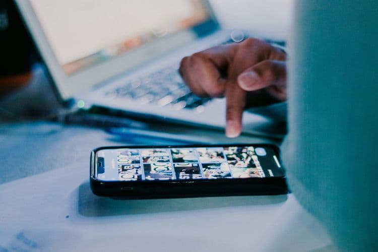Eine Person scrollt auf einem Smartphone durch einen bildbasierten Feed, im Hintergrund ist ein Laptop zu sehen.