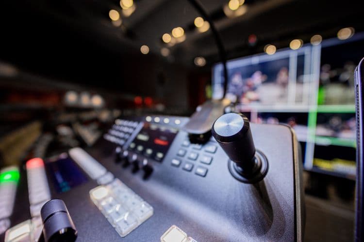 Nahaufnahme eines technischen Bedienpults mit Steuerhebeln und unscharfen Monitoren in einer Fernsehregie.