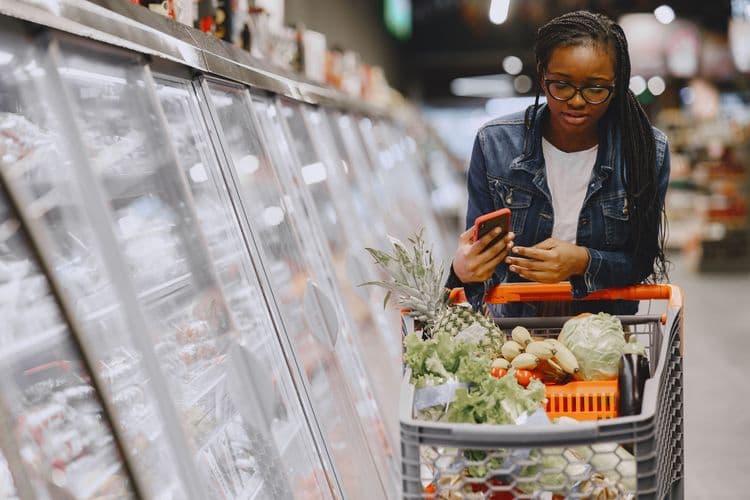 Kundin mit Smartphone und gefülltem Einkaufswagen in einem Supermarkt.