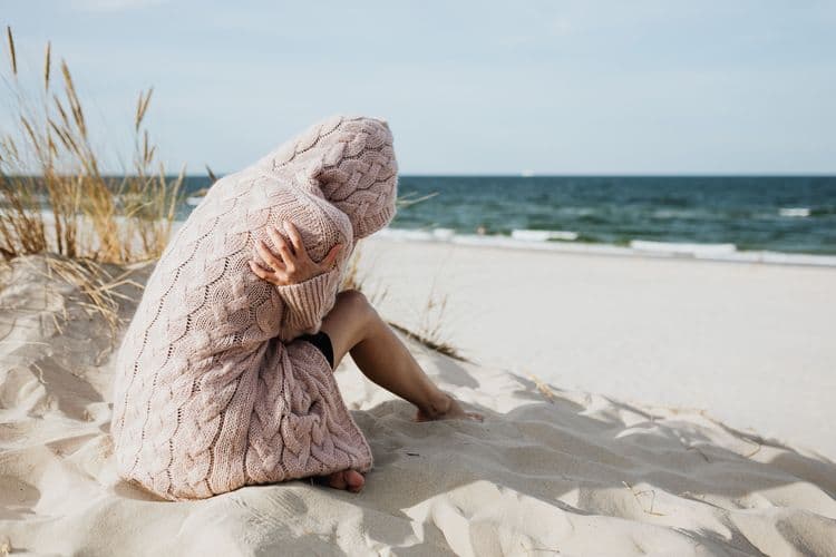 Person sitzt in eine Decke gehüllt am Strand und blickt aufs Meer.