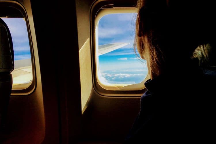Person sitzt im Flugzeug am Fenster und blickt auf Tragfläche und Himmel.