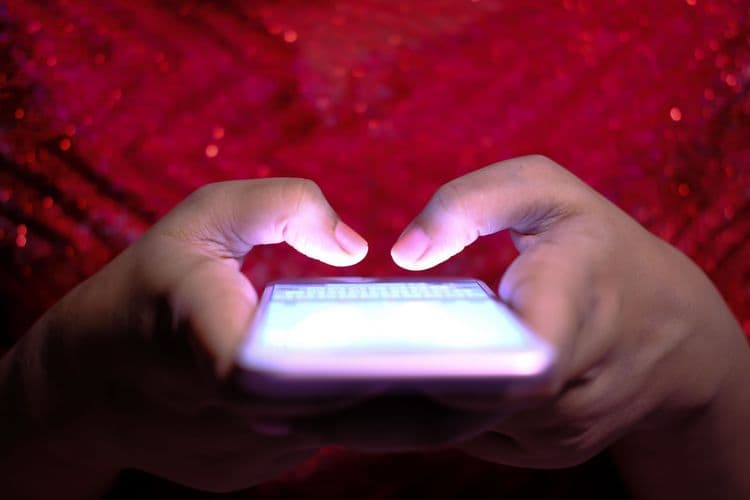 Hände bedienen ein leuchtendes Smartphone vor rotem Hintergrund.