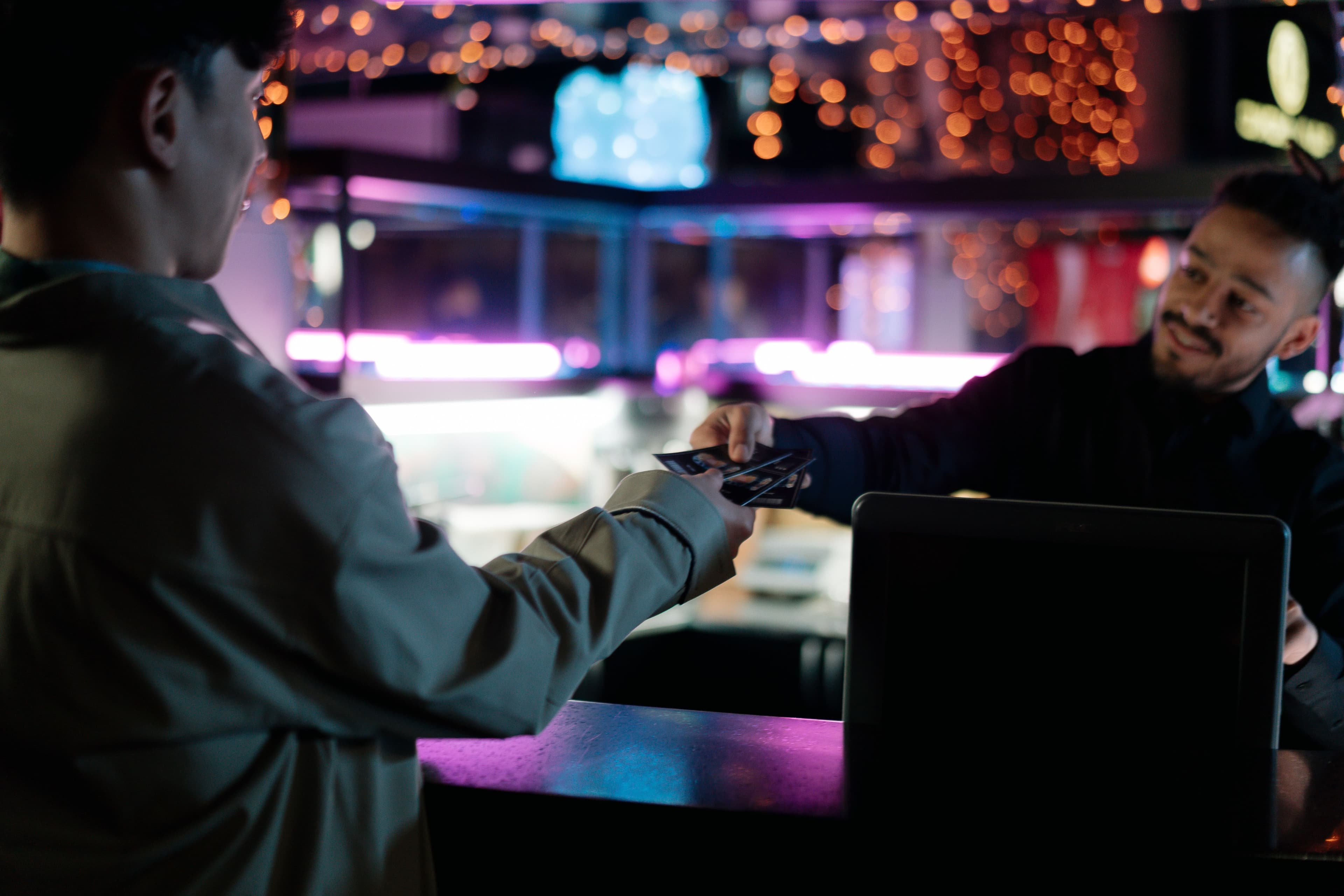 Zwei Personen übergeben in einer Bar bei Neonlicht Karten an einem Tresen mit Laptop.