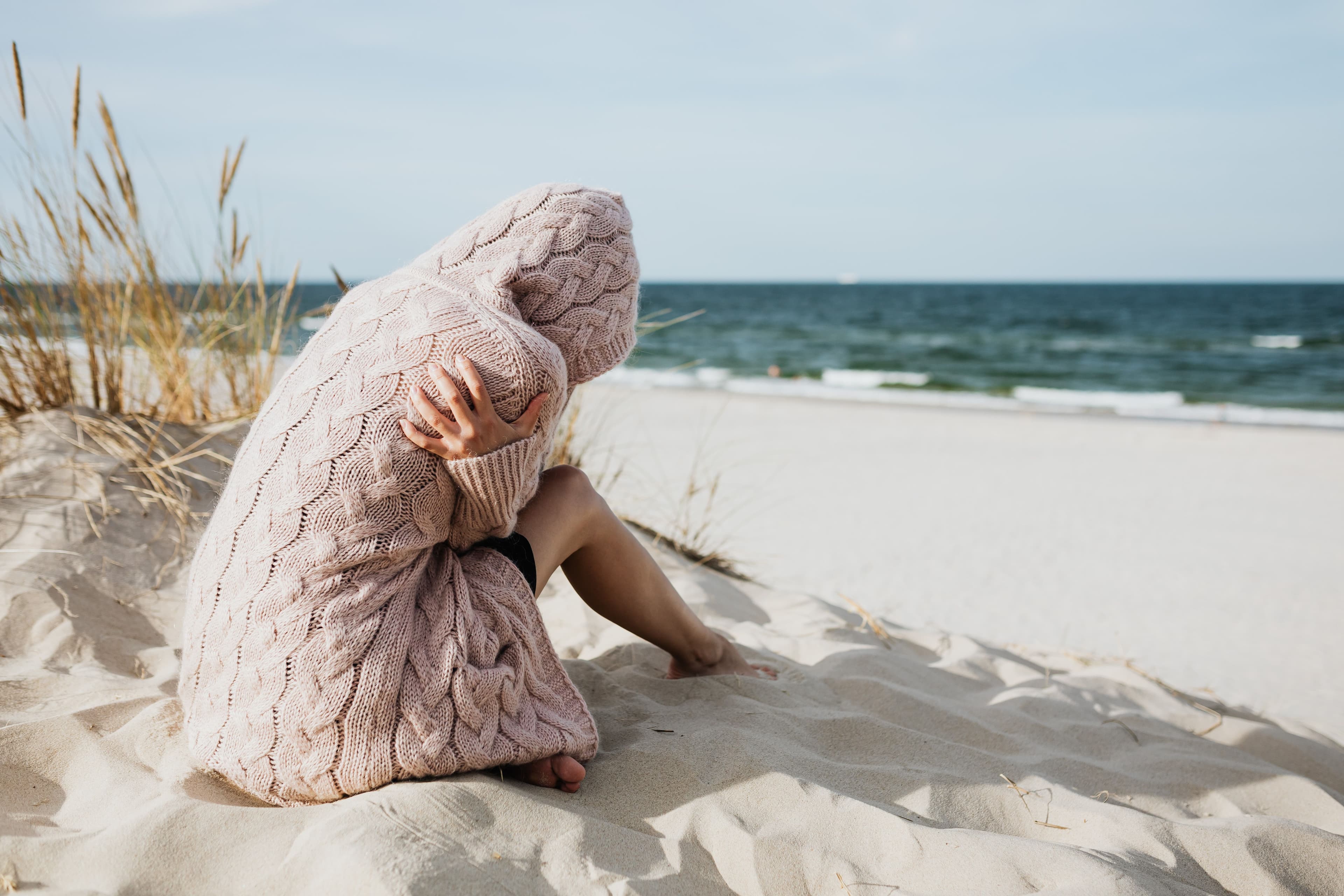 Person sitzt in eine Decke gehüllt am Strand und blickt aufs Meer.