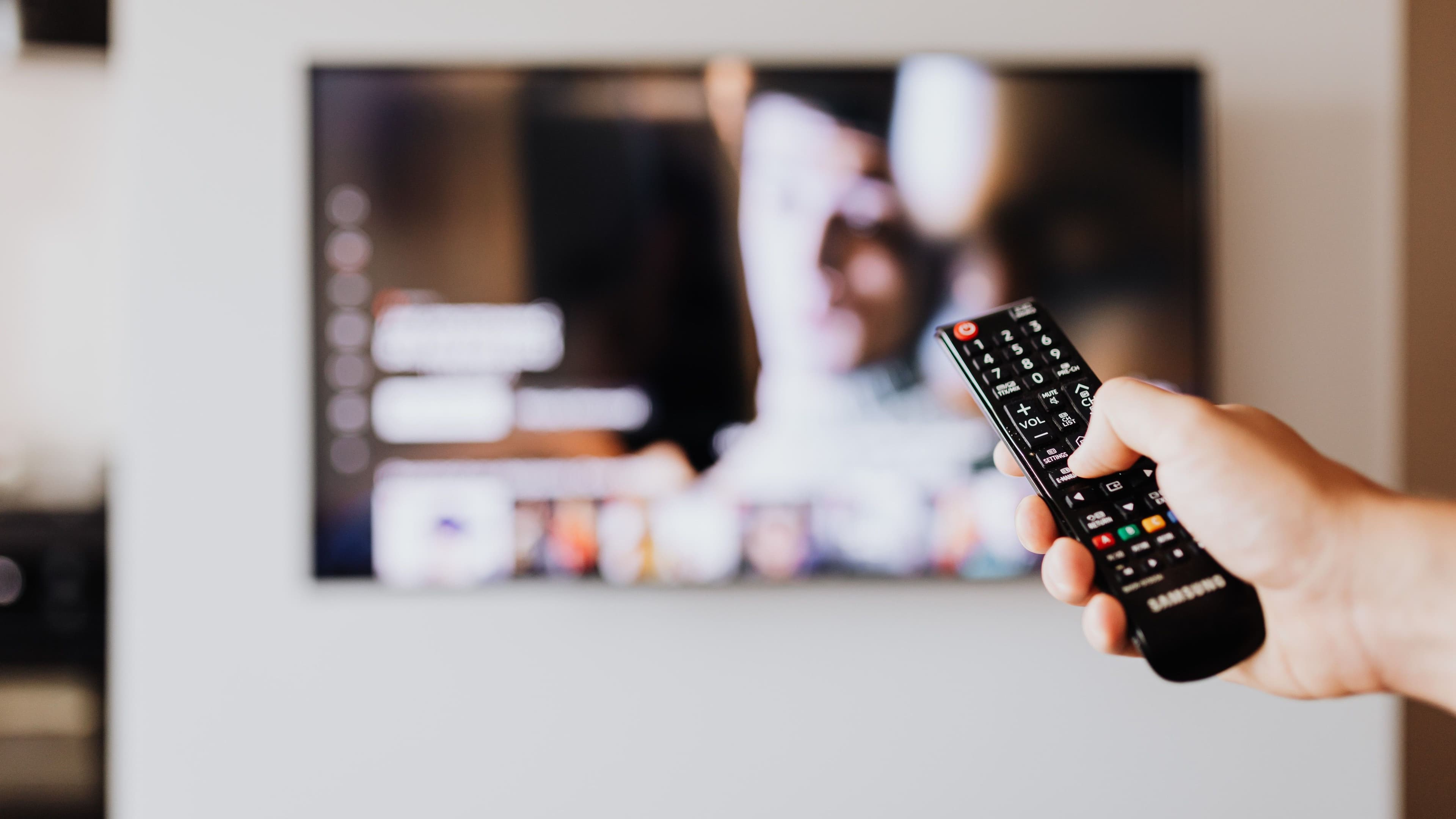 Hand mit Fernbedienung vor einem unscharf dargestellten Fernseher.