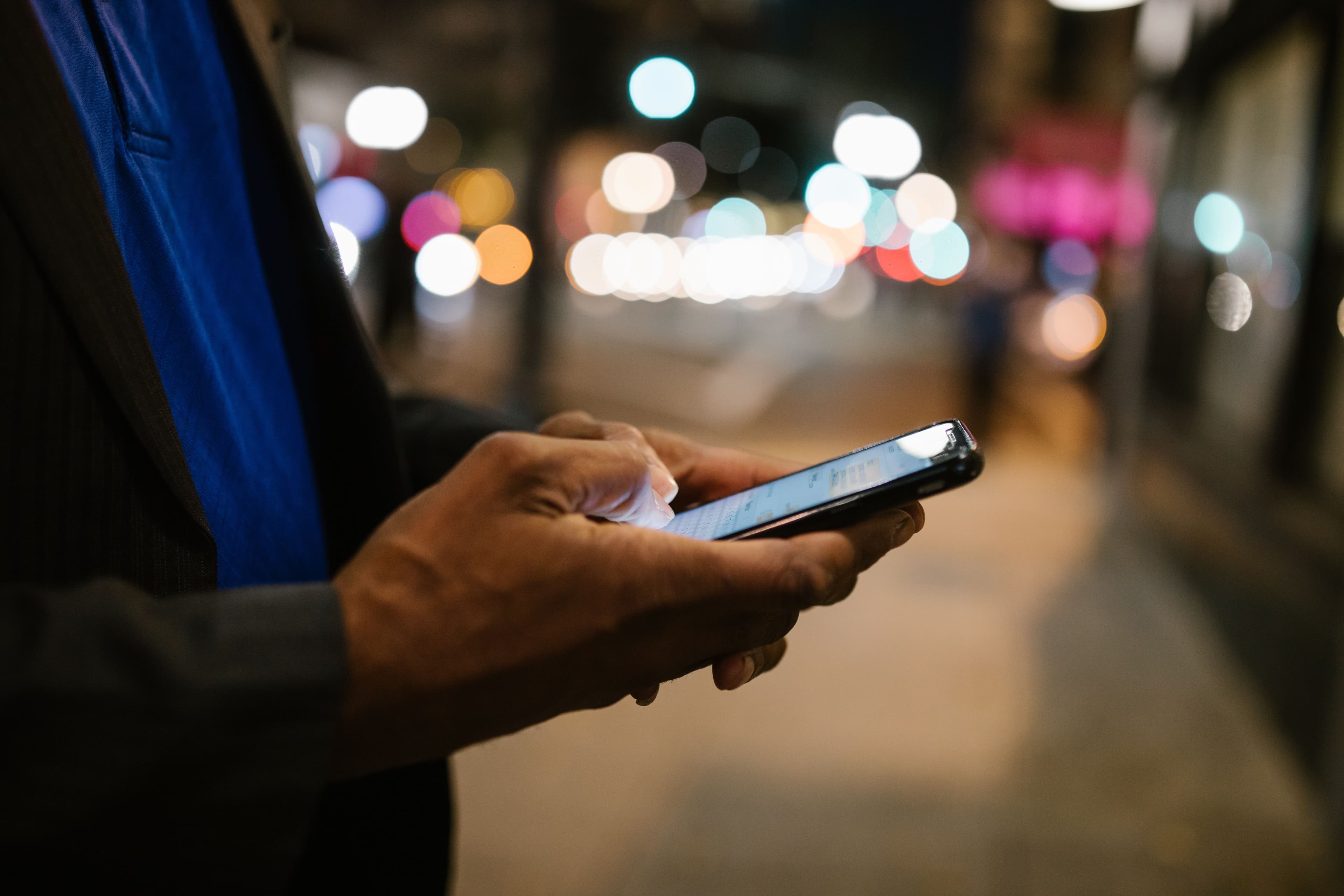 Eine Person nutzt abends ein Smartphone vor unscharfen Lichtern im Hintergrund.
