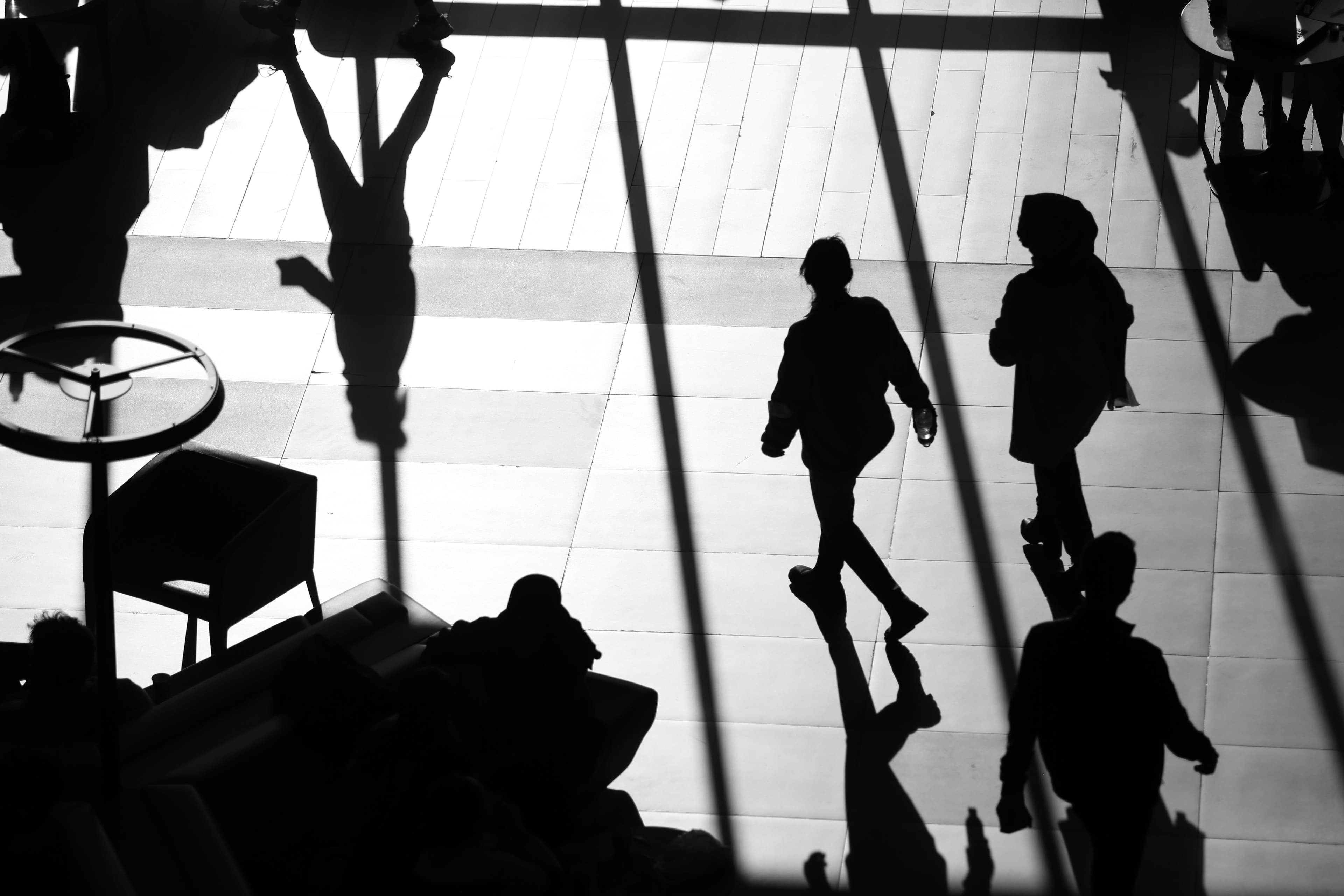 Schwarz-Weiß-Aufnahme von Menschen, die in einem hellen Innenraum als Silhouetten unterwegs sind.