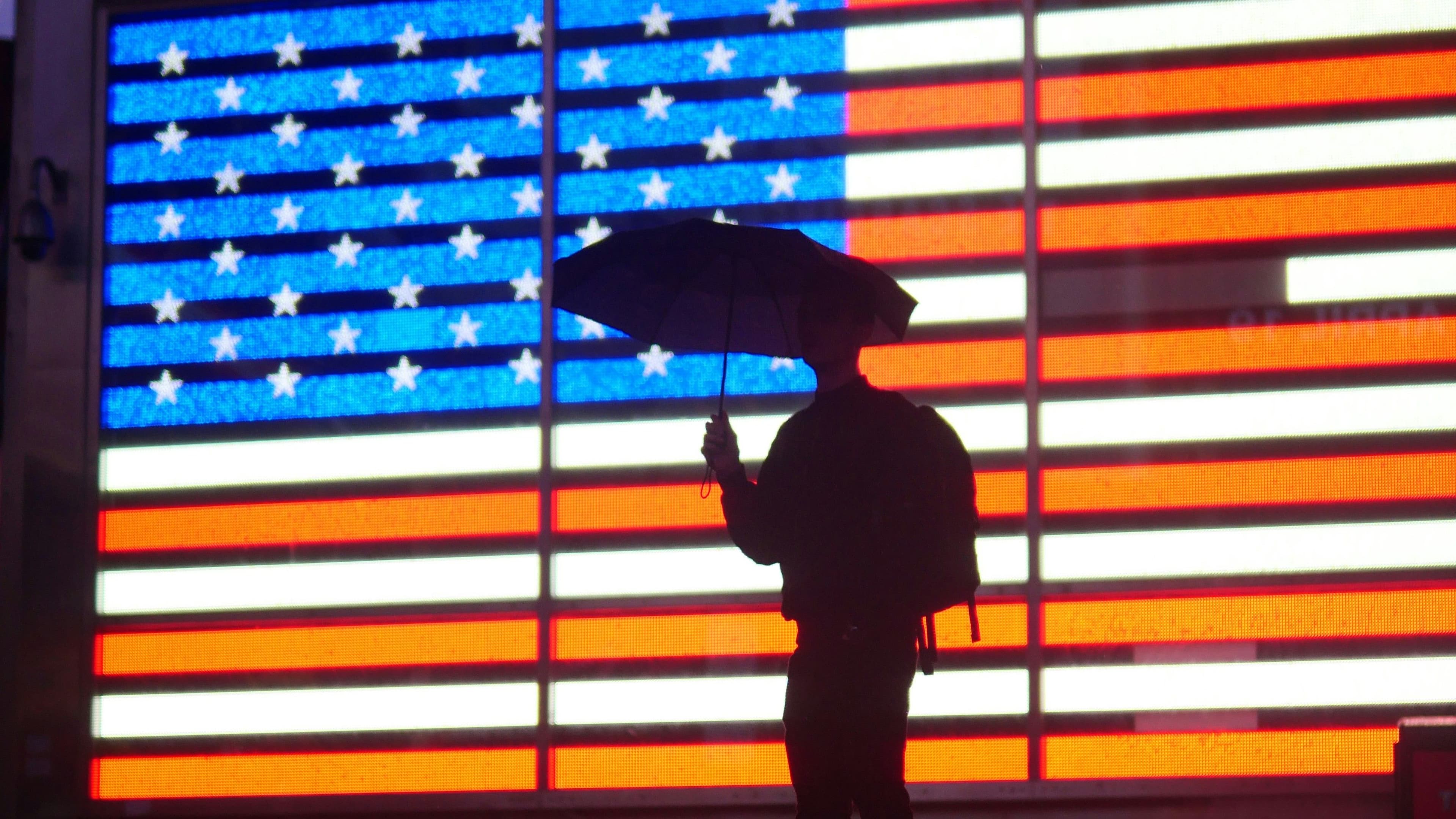 Eine Person mit Regenschirm steht vor einer hell leuchtenden digitalen US-Flagge.