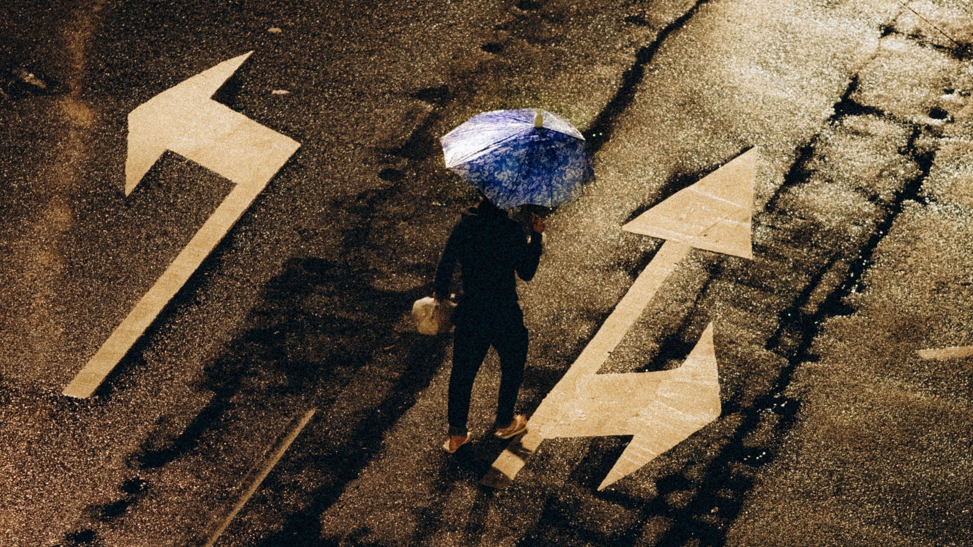 Person mit Regenschirm steht zwischen mehreren Richtungspfeilen auf einer nassen Straße.