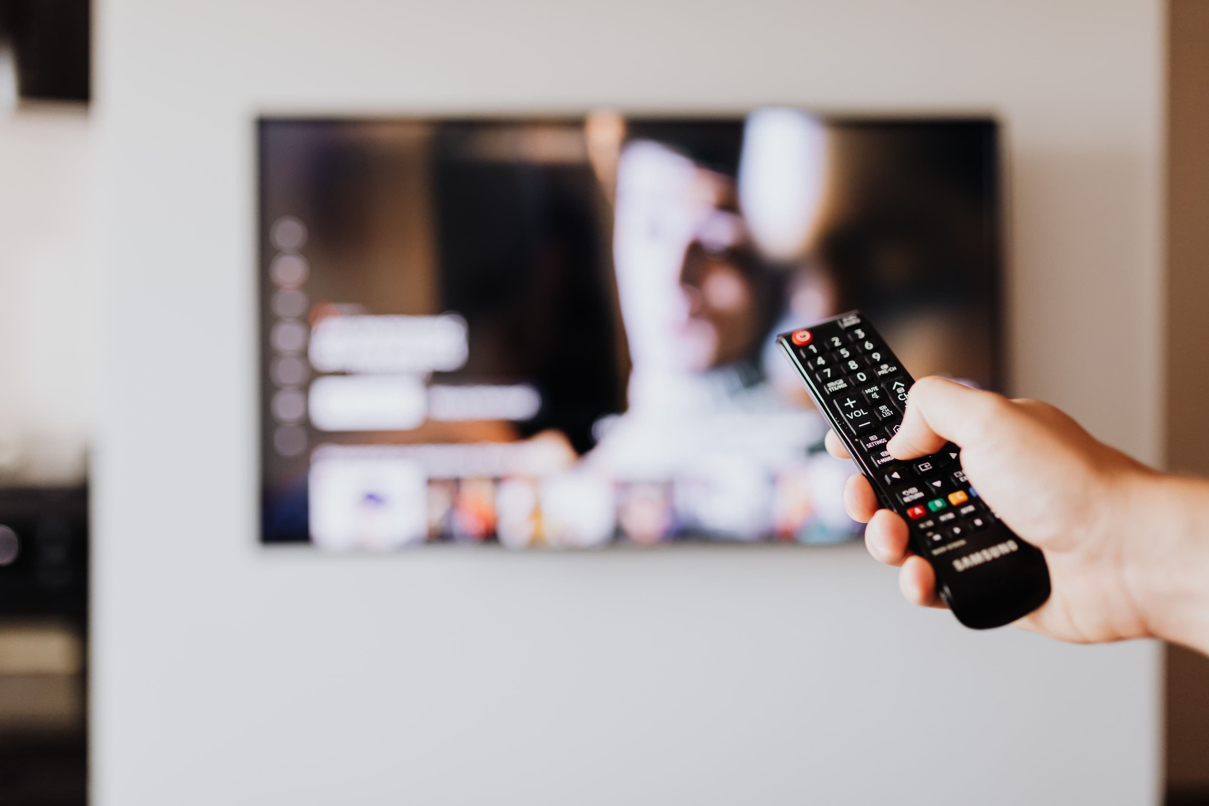 Hand hält eine Fernbedienung vor einem Fernseher mit unscharf dargestellter Streamingoberfläche.
