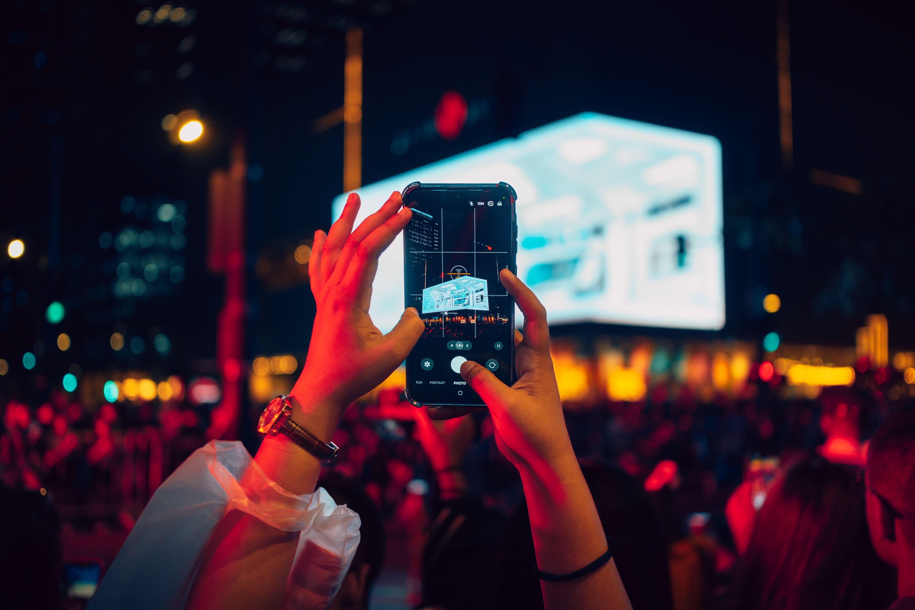 Eine Person fotografiert mit dem Smartphone eine große leuchtende Fläche im öffentlichen Raum bei Nacht.