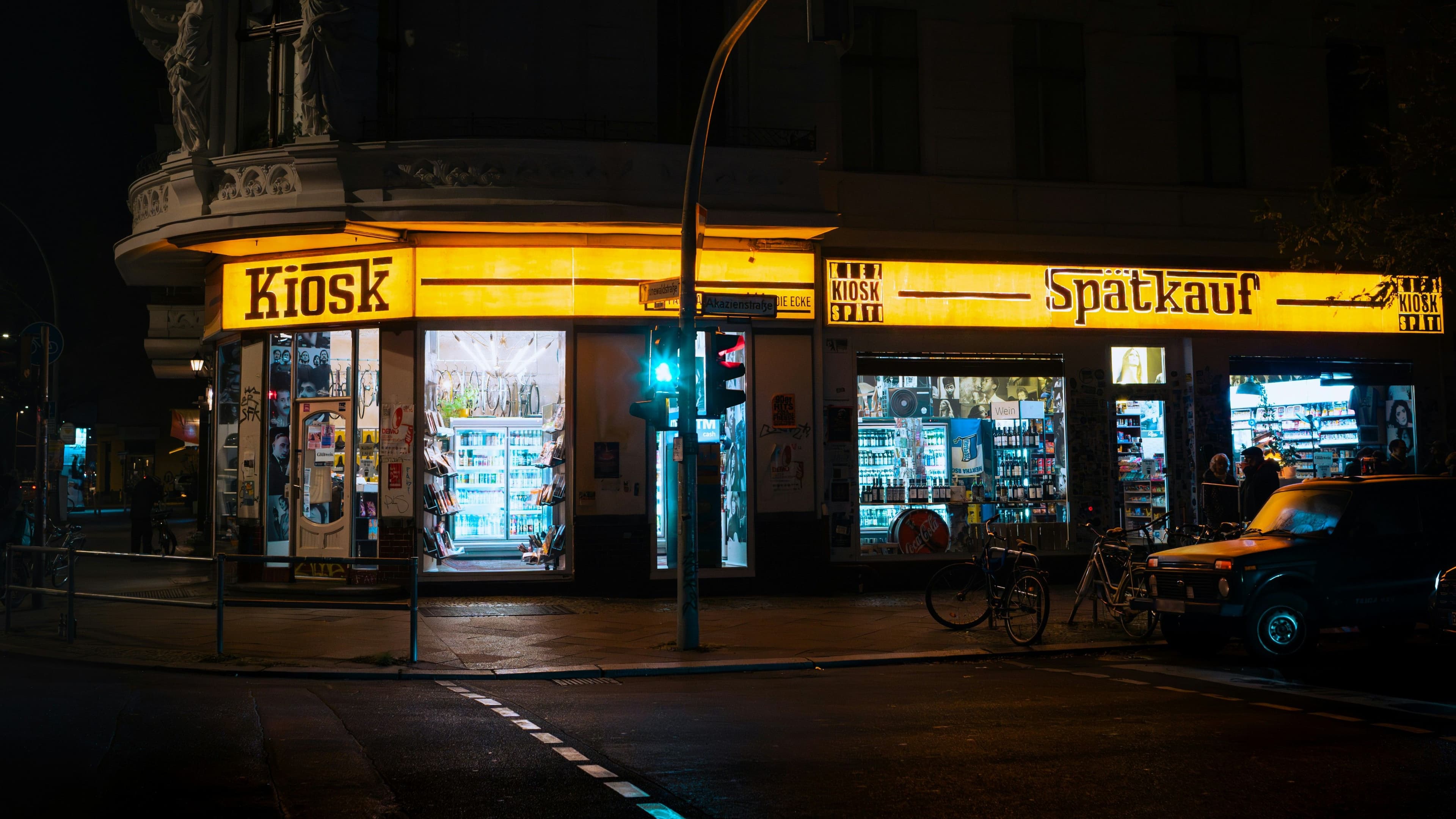 Nächtliche Außenansicht eines beleuchteten Berliner Spätkaufs mit Kiosk-Schriftzug, Schaufenstern, Getränkeregalen und Fahrrädern vor dem Laden.